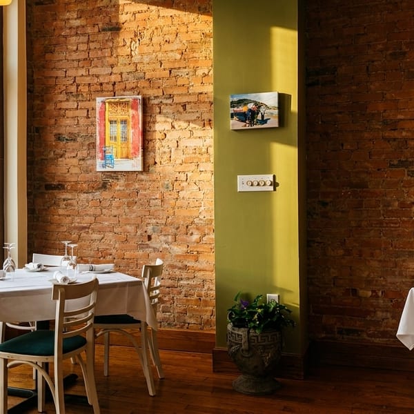 Sunlight streams into a restaurant dining room with brick walls, set tables, and street view.
