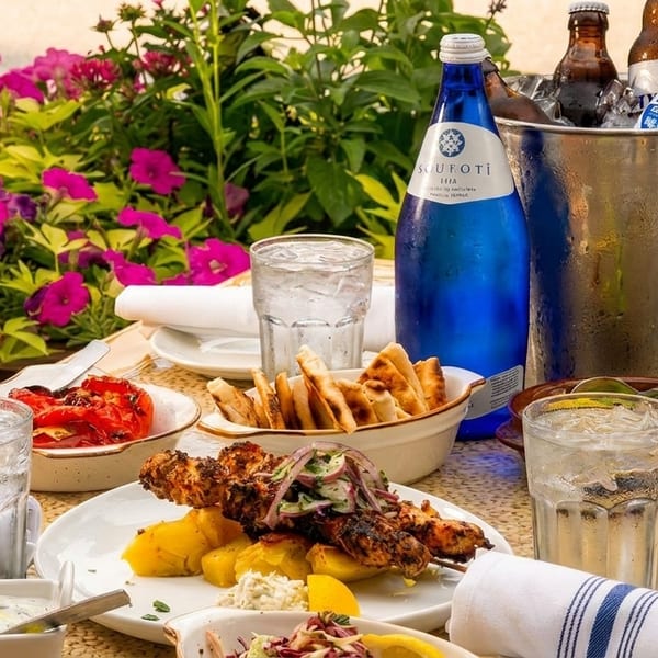 A vibrant outdoor dining table with Greek food, drinks, and pink flowers in the foreground.